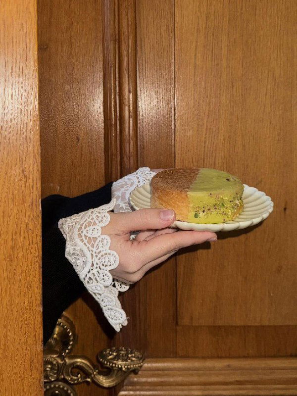 Рука держит тарелку с кусочком свежей выпечки от «Футуры»: кружевная манжетка на рукаве, деревянный фон и золотистая корочка десерта.