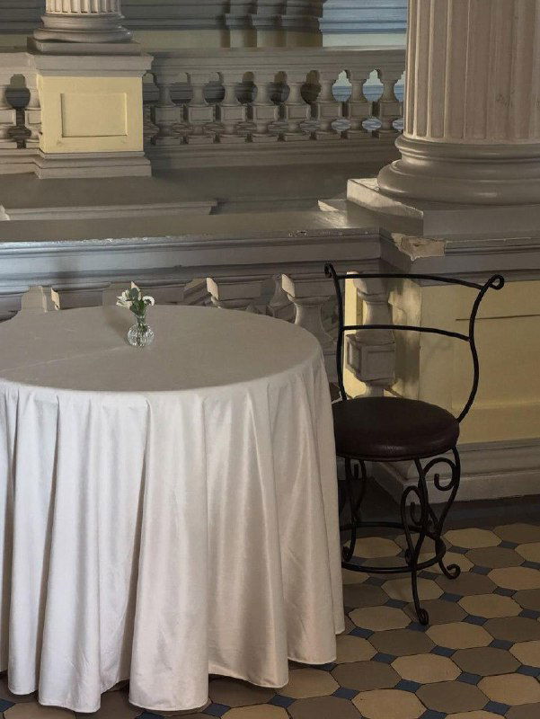 Крупный план круглого столика в Кофейной комнате: скатерть до пола, кованый стул и небольшая вазочка с цветком на фоне колонн.