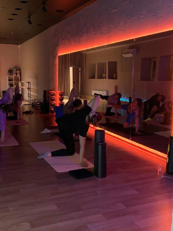 Групповая тренировка в затемнённом зале: люди на ковриках делают растяжку или пилатес на фоне подсветки и зеркала — уютная студия занятия.