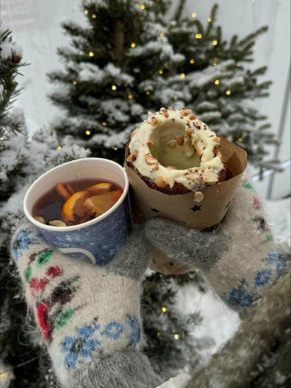 Рука в тёплой варежке держит чашку горячего напитка и рядом чешский трдельник на фоне заснеженной ёлки и праздничных огней