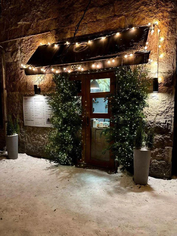 Деревянная дверь и вывеска Cozy Coffee украшены гирляндами и огоньками, каменная стена и снег подчёркивают атмосферу маленького уютного кафе