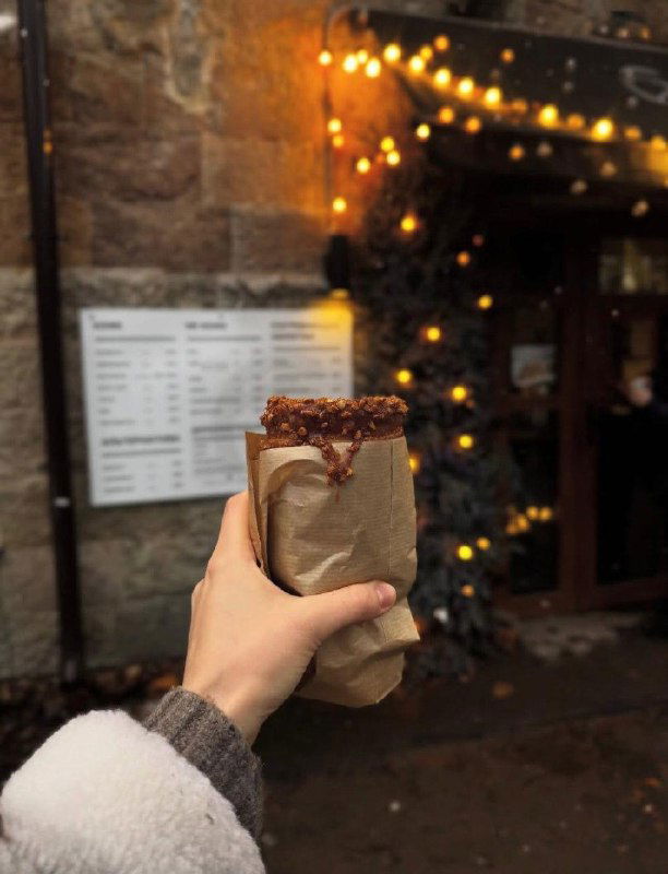 Трдельник в бумажной обёртке держат на переднем плане, на заднем плане размыто меню кофейни и тёплое освещение гирлянд