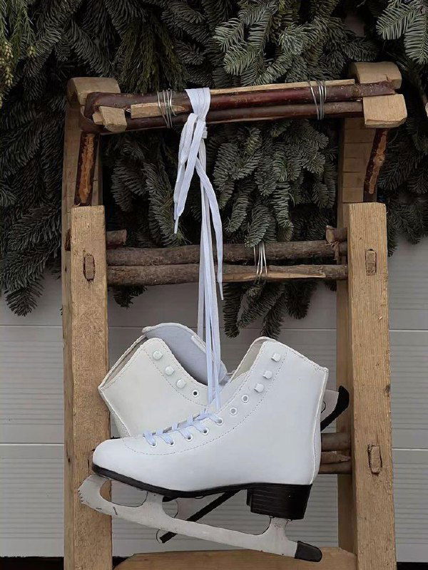 Белые фигурные коньки, подвешенные на деревянной подставке на фоне хвойных ветвей — деталь зимней ярмарки или пункта проката на катке.