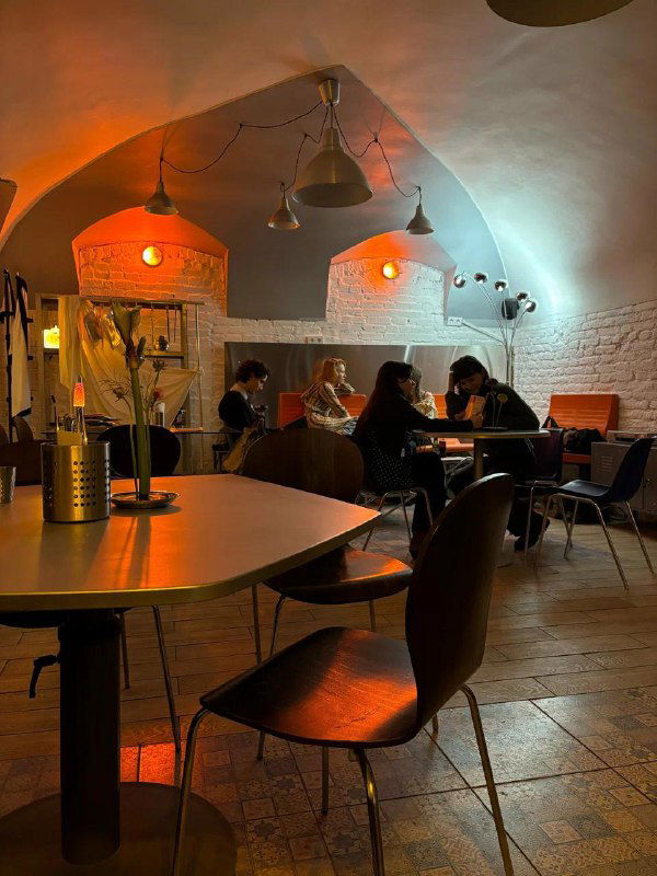 Кафе-зона с арочными потолками, столиками и посетителями, подсвеченная тёплым оранжевым светом — уютное пространство на Ваське.