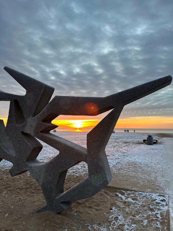 Побережье на закате с абстрактной скульптурой на пляже, холодный воздух и лёгкая изморозь на береговой линии.