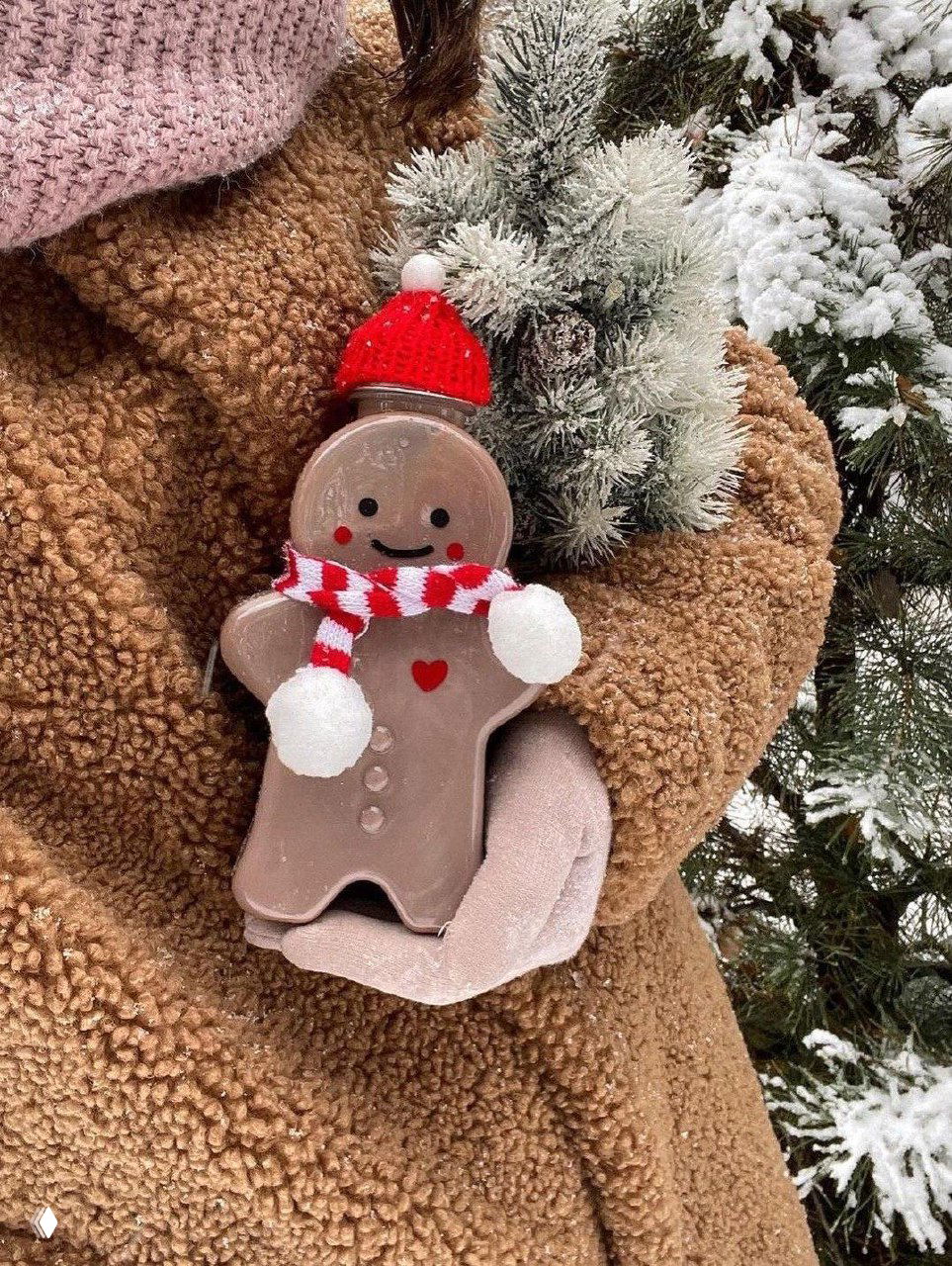 Многоразовая бутылочка‑имбирный человечек в красной шапке держится в руках в коричневом пальто на фоне снежной ёлки.