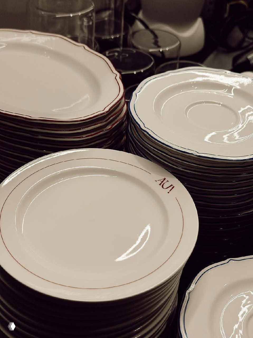 Стопка белых тарелок с тонкой каймой, одна тарелка с подписью Au Delà на переднем плане — аккуратная сервировка посуды.