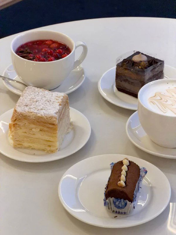 На столе в кафе Север‑Метрополь: чашки с латте и ягодным напитком, рядом десерты — Наполеон, шоколадный кусок и порция компота.