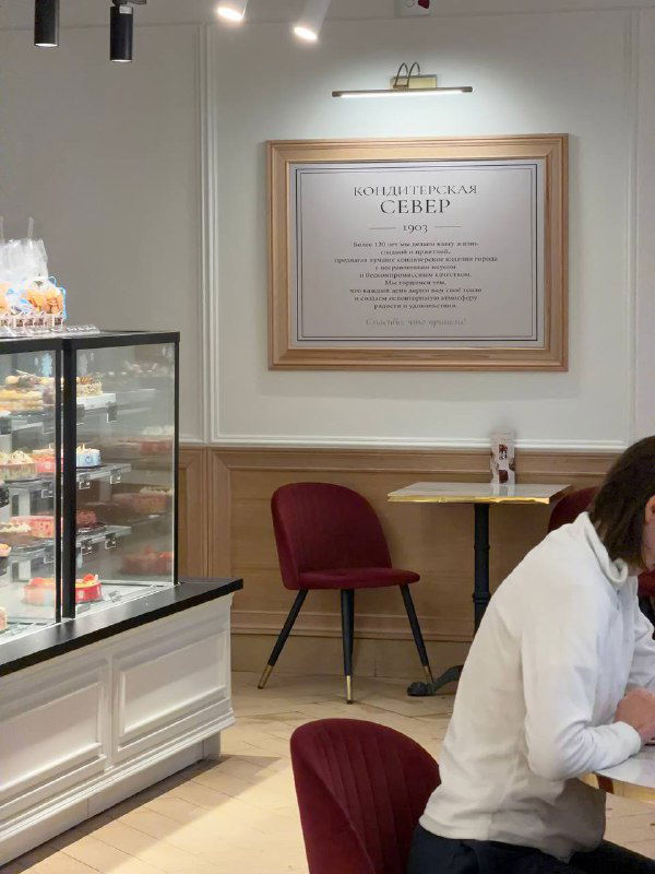 Интерьер кондитерской Север‑Метрополь: витрина с десертами, уютные столики и стулья, на стене оформленная вывеска с текстом истории.