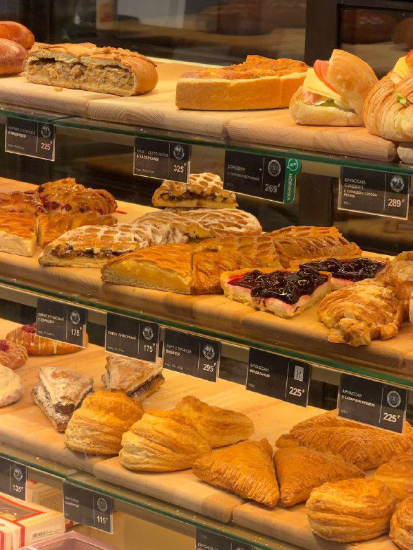Полка с выпечкой и слоёными изделиями в витрине Север‑Метрополь: круассаны, пироги и пирожные с ценниками в Петербургской пекарне.