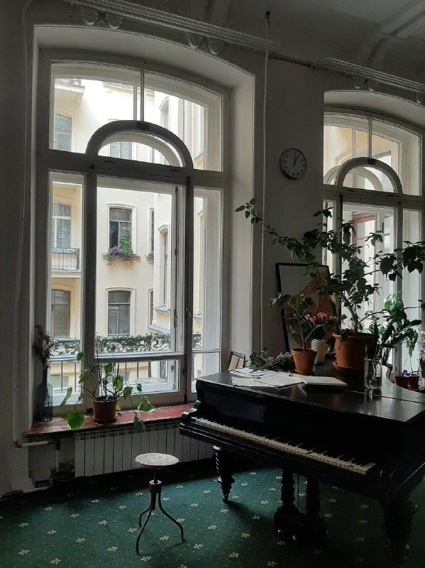 Рояль у больших арочных окон в комнате Дома Бака, на подоконнике комнатные растения и вид на внутренний двор и фасады зданий.