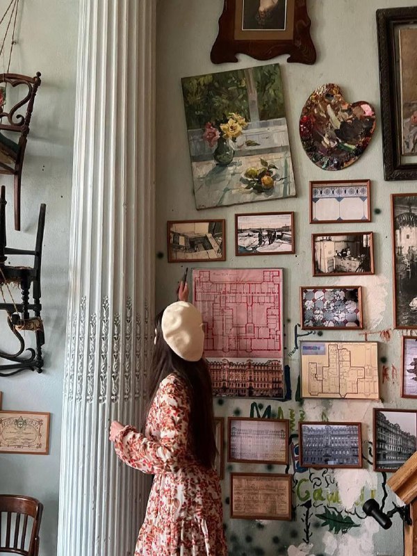 Манекен в винтажном платье возле колонны; на стене — множество рам и картин, создающих музейную атмосферу интерьера.