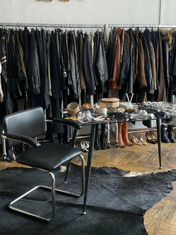 Интерьер секонд‑шопа: ряд кожаных курток и пальто на вешалках, перед ними стол с мелкими вещами и стул — атмосферный кадр магазина.