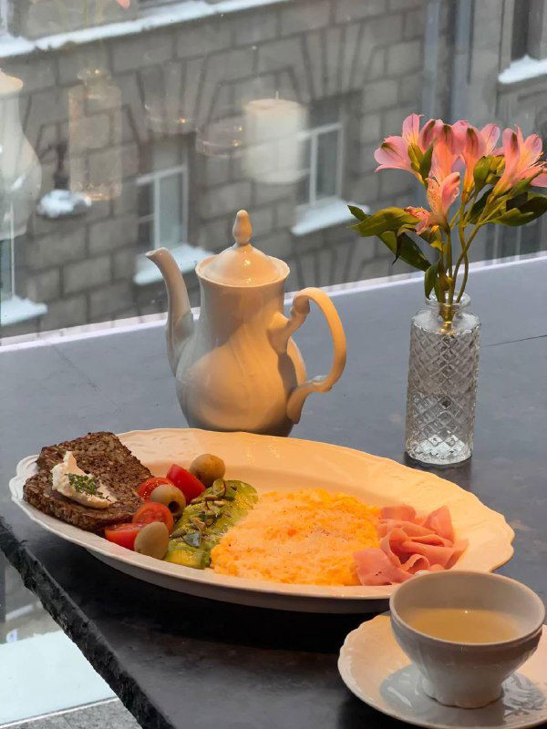 Большой завтрак TETTO: омлет, тамбовский окорок, маслины и овощи на тарелке рядом с чайником на подоконнике с городским видом и аккуратной сервировкой.