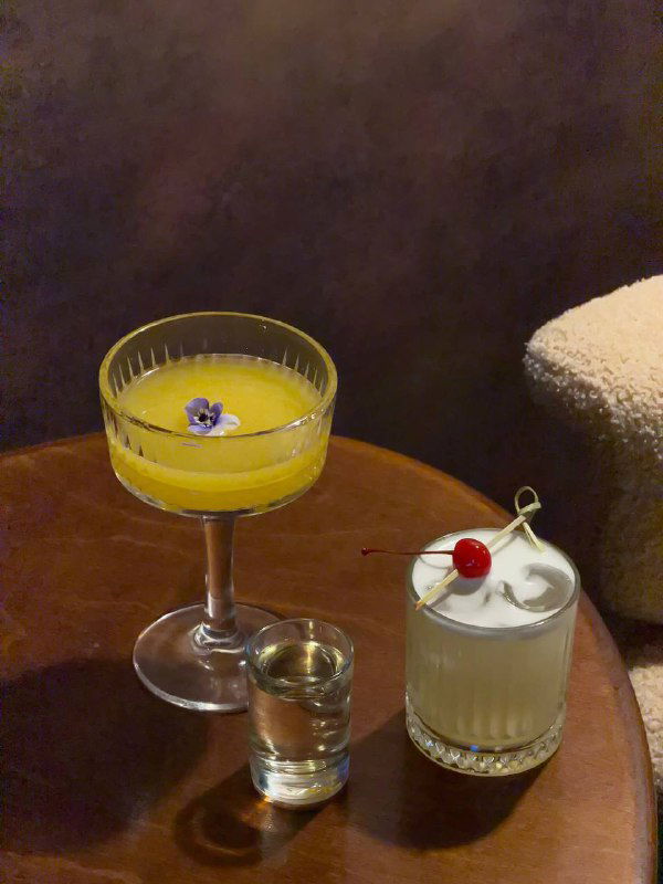 Два коктейля на низком столике: желтый coupe-коктейль с цветком и кремовый коктейль с вишенкой; рядом рюмка для шота — подача напитков в баре.