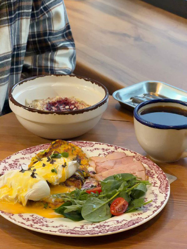 Завтрак в 8:am: тарелка с омлетом и лососем, творожный сыр и микс-салат, рядом миска овсянки и чашка чёрного кофе на деревянном столе в бистро.