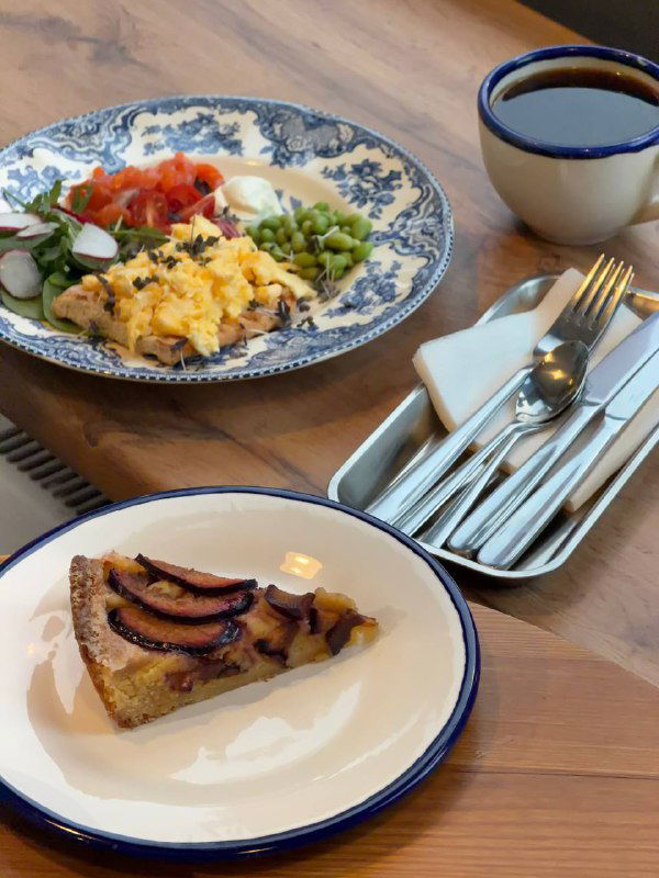 Несколько блюд на столе в кафе: скрэмбл, эдамаме и салат, на переднем плане сливовый тарт на белой тарелке — домашние десерты 8:am.