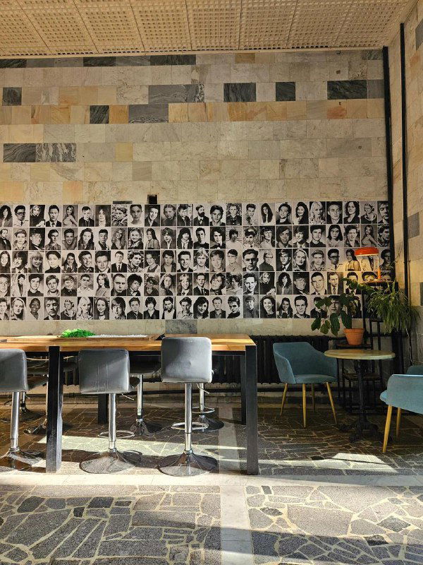 Внутренний зал кофейни с большой стеной, украшенной ретро-фотопортретами; столы и стулья на фоне индустриального декора.