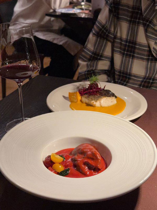 Две тарелки на столе: в одной сочный кусок рыбы с соусом, в другой — насыщенно-красное блюдо, рядом бокал белого вина и чашки для чая