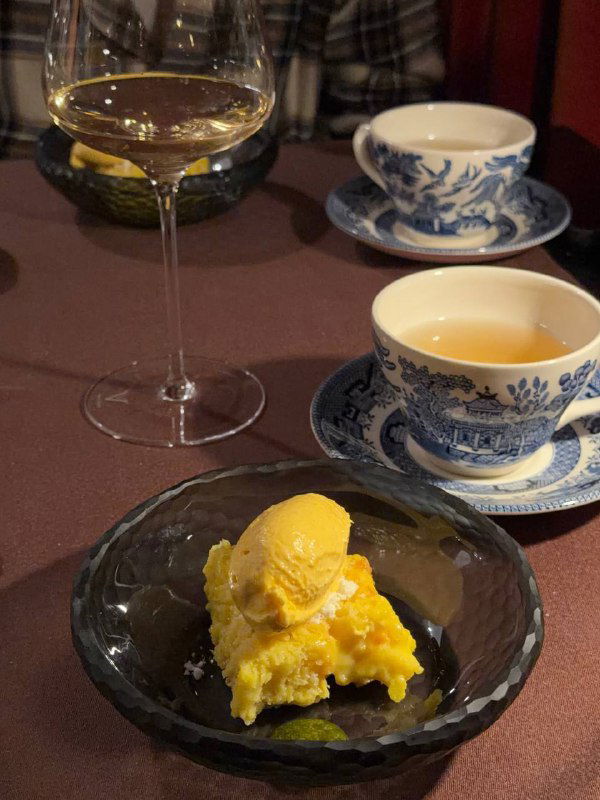 Небольшая порция яркого желтого десерта в чёрной пиале, рядом чашка с чаем и бокал белого вина — финал сета в ресторане ЕМ