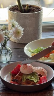 Вафля с кремом из чоризо и яйцом пашот: крупный план блюда на деревянном столе, рядом нож и цветы на подоконнике.