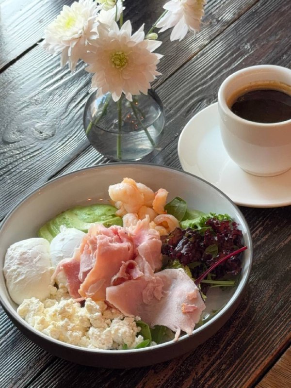 Крупный план второго варианта завтрака: тарелка с креветками, рикоттой, окороком и зеленью, чашка кофе и светлое деревянное покрытие стола.