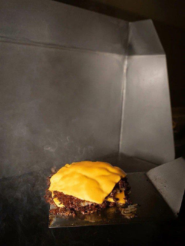 Процесс приготовления смэш-бургера: прижатая котлета на лопатке/гриле под кусочком плавленого сыра, кадр из кухни заведения.