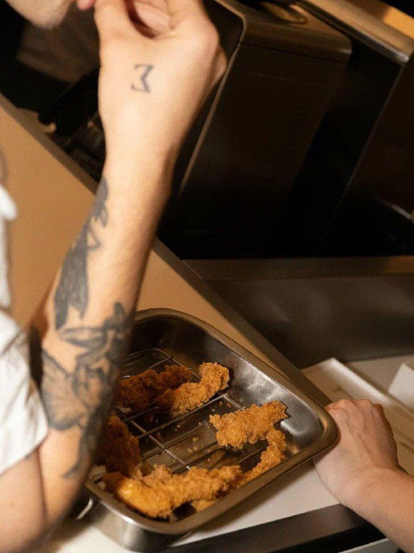 Кадр из кухни BUMO: руки повара с татуировками держат лоток с панированными кусочками, вид технологического процесса и кухни.