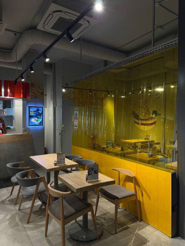 Интерьер рамэн-бара Asiatiq: светлые деревянные столы и стулья, отделение с жёлтыми шторками и минималистичный дизайн помещения.