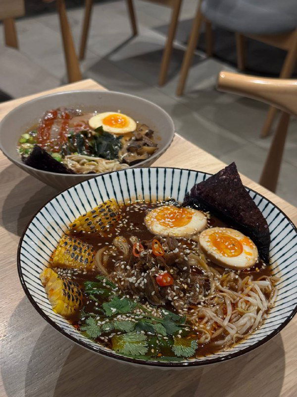 Две миски рамэна на столе: в переднем плане яркий бульон, яйцо, нори и зелень, в глубине ещё одна порция — аппетитный вид блюда.