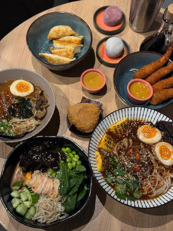 Несколько тарелок с разными блюдами: мисо-рамэн, онигири, гедза и креветки на общем столе, вид сверху на набор заказанных позиций.