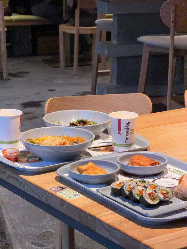 Подносы с рамёном и закусками на столе у окна: две миски супа, роллы и пого́ди в лотках в формате быстрого обслуживания.