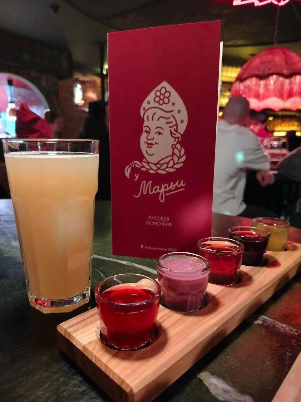 Дегустационная подача настоек: поднос с пятью рюмками разных настоек, рядом высокий коктейль и красное меню «Марья» на барной стойке, крупный план.