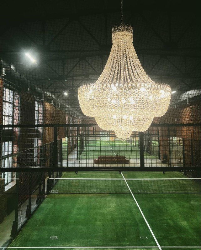 Вид с трибун на корт клуба «Ракета»: высокие потолки, кирпичные стены и сияющая хрустальная люстра над игровым пространством с искусственным покрытием.