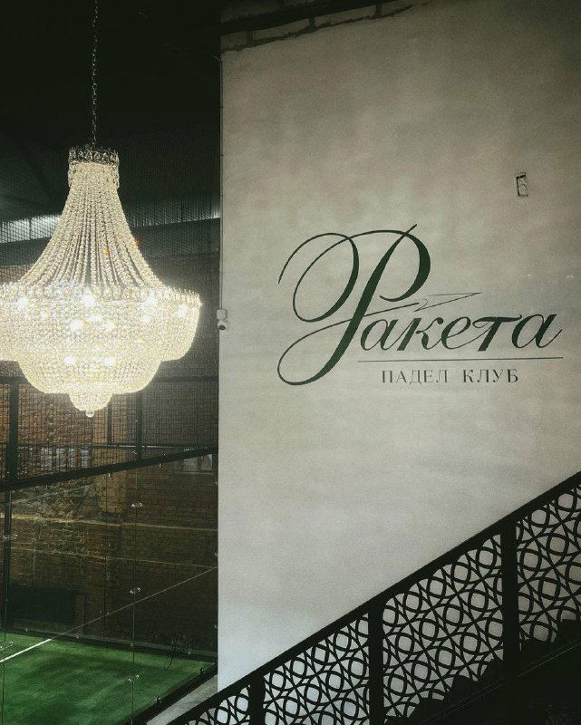 Логотип и брендирование клуба «Ракета» на стене рядом с падел‑кортом: крупный надписьный знак на фоне белой стены и лестничного ограждения в интерьере.