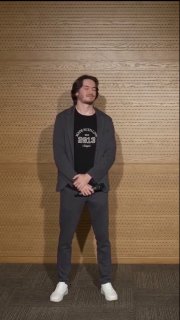 Человек в тёмной одежде стоит в полный рост перед перфорированной деревянной стеной — фото с презентации на мероприятии.