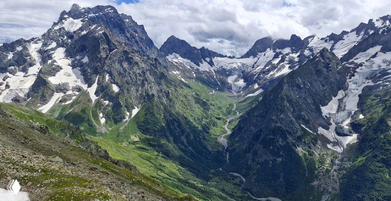 Широкий горный вид с крутыми скалами и глубокими цирками, характерный для окрестностей Домбая с облачным небом над вершинами.