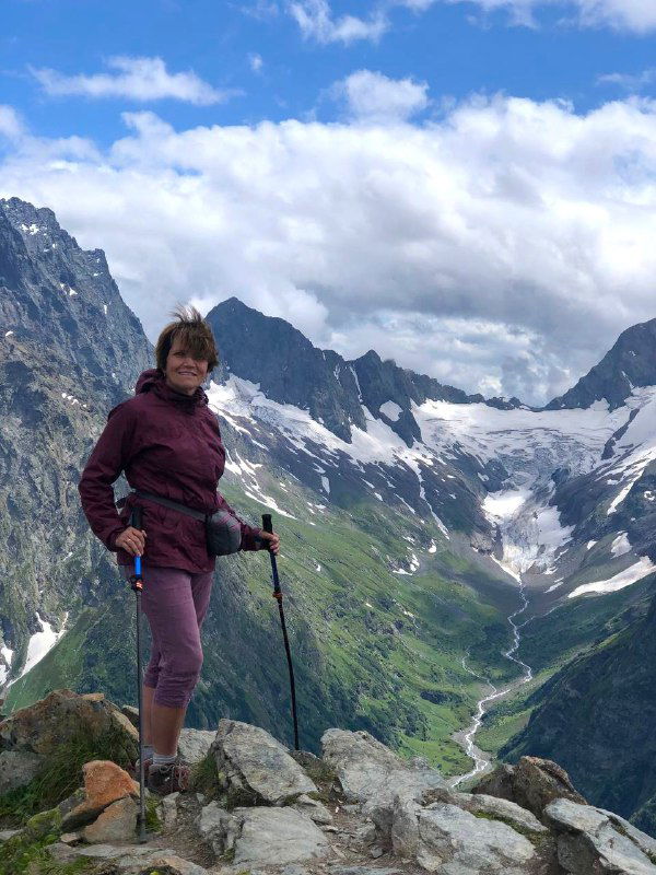 Автор поста стоит на камнях с треккинговыми палками, позади видна глубокая долина и горные пики — снимок с личного маршрута в Домбае.