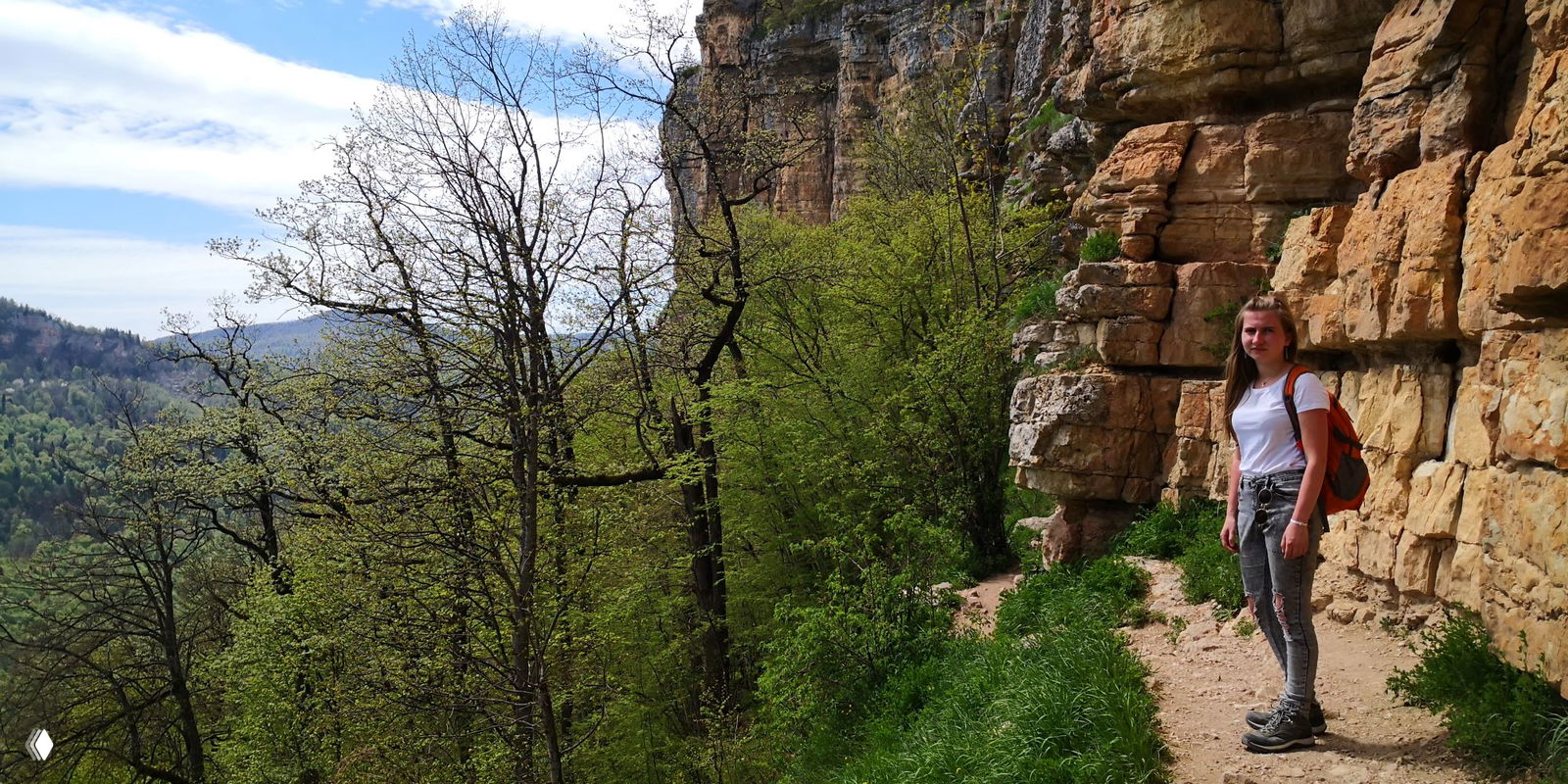 Туристка идёт по узкой тропе у скальной стены, справа видны мощные желтоватые скалы, слева простирается лесистый склон и панорама ущелья.