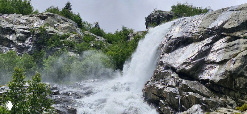 Крупный план Алибекского водопада: бурные струи воды падают по скалам, образуя брызги и шумное каменистое русло внизу