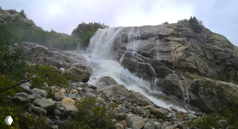 Алибекский водопад с нижней точки съёмки: каскад воды спускается по уступам скал, вокруг камни и редкая растительность