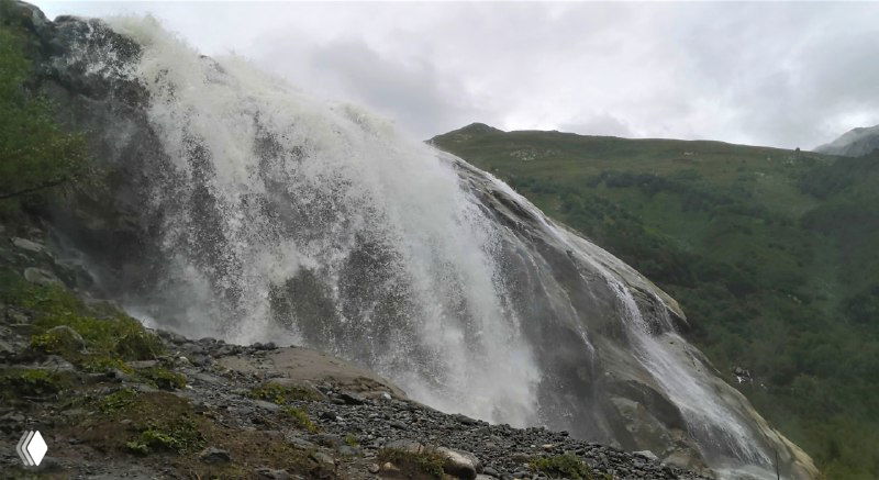 Широкий каскад водопада в ущелье: плотный поток воды по склону, мокрые камни внизу и зелёные склоны вокруг