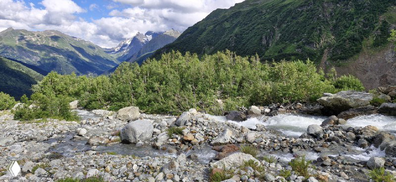Речная долина с галечным руслом в Алибекском ущелье: быстрый поток между камнями, кустарники вдоль берегов и вид на окрестные склоны