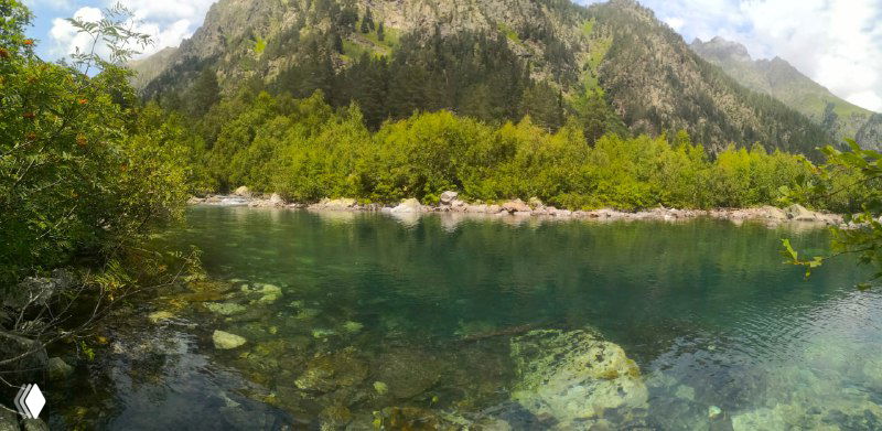 Широкий панорамный снимок озера и береговой растительности, зелёные оттенки воды на фоне горной стены и хвойных лесов.