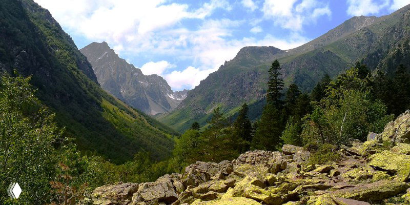 Долина с курумником и хвойным лесом, тропа идёт вверх между камнями к более высоким озёрам в скалистой части Домбая.