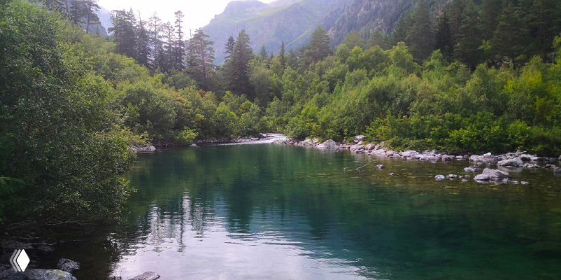 Зелёное горное озеро с отражением деревьев и неба, каменистый берег и густая прибрежная растительность в долине Бадука.
