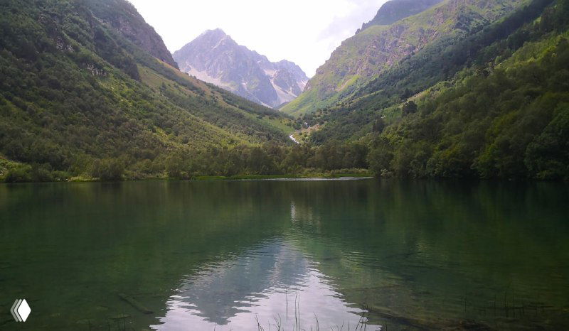 Широкий вид на озеро с отражающимися вершинами гор, узкая V-образная долина и лесистые склоны по обе стороны водоёма.