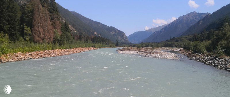 Река Теберда в каменистом русле перед подвесным мостом, мутноватая горная вода, галька на берегах и горы на горизонте.