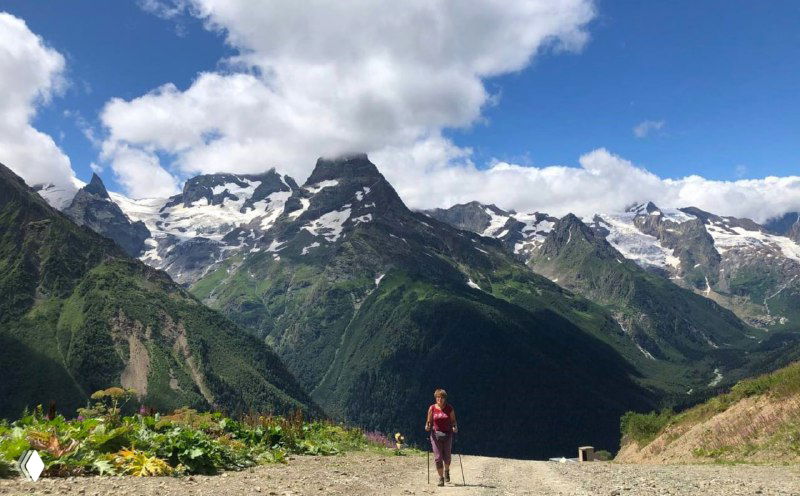 Треккер идёт по тропе на фоне массивных гор и голубого неба — видна часть подъёма от одной из станций канатки.