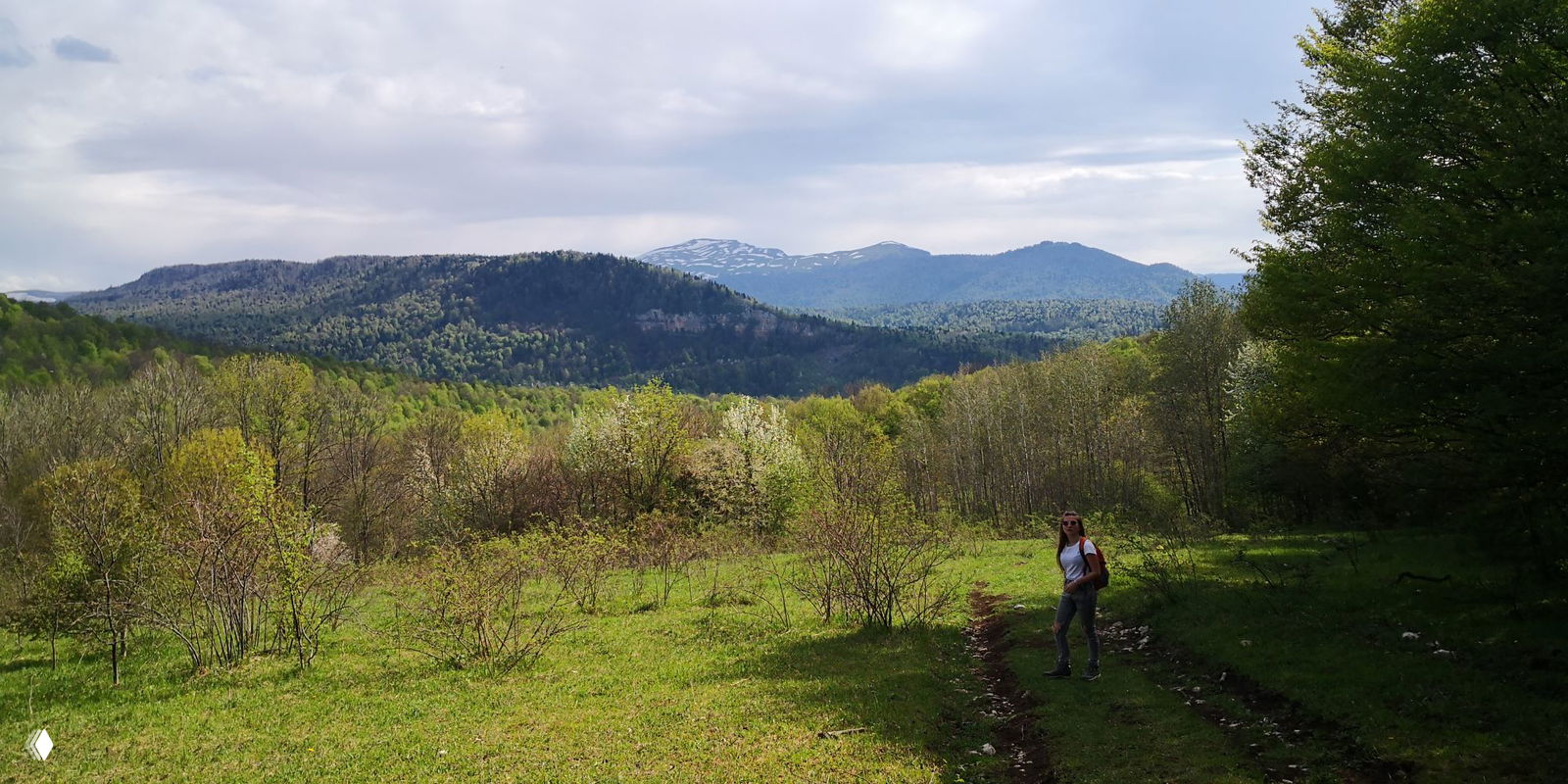 Туристка идёт по лесной поляне и тропе в весеннем лесу, видны удалённые горные силуэты и зелёные ковры на подступах к плато Лагонаки.